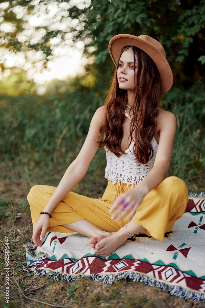 Fototapeta premium Woman in ecological clothing in a hippie look sits on a colored plaid meditating in the lotus position smiling and looking at the autumn sunset in the park