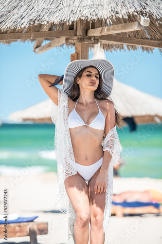 Beautiful brunette girl in a white bathing suit.Portrait of professional swimwear model posing provocatively outdoor.Smiling sensual lady with long legs posing in white bikini and hat the beach.