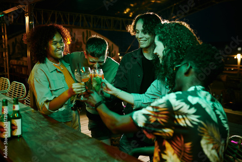 Group of joyful intercultural friends clinking with glasses of cocktails while standing by bar counter and enjoying party in night club