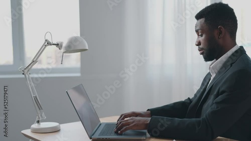 An African-American businessman in a business suit works in his office and looks at his laptop monitor with incomprehension and surprise