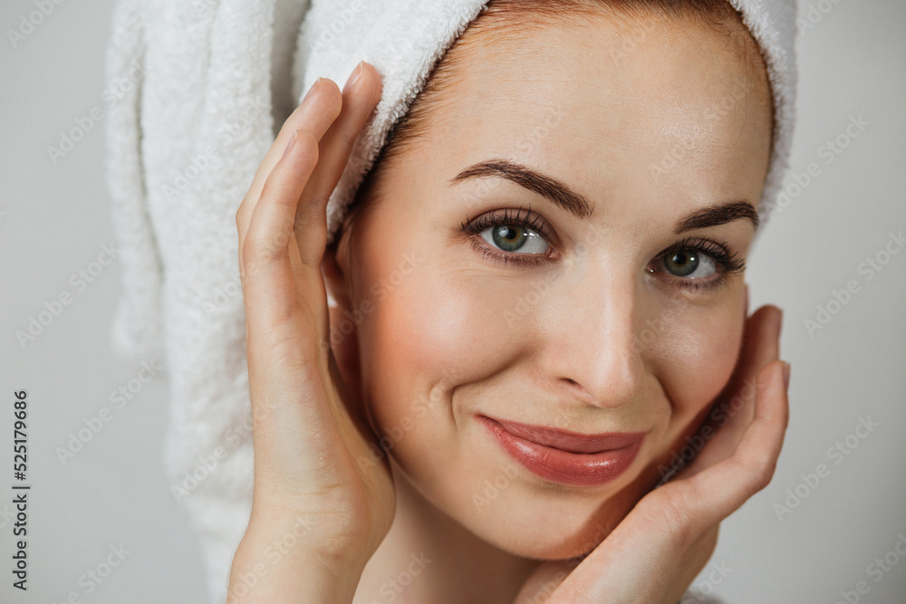 Portrait of smiling caucasian woman with healthy skin in towel posing over grey background. Happy young lady touching her perfect face with hands. Portrait of smiling caucasian woman with healthy skin in towel posing over grey background. Happy young lady touching her perfect face with hands.