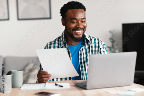 Canvas Print Cheerful African American Freelancer Man Using Laptop Holding Papers Indoor
