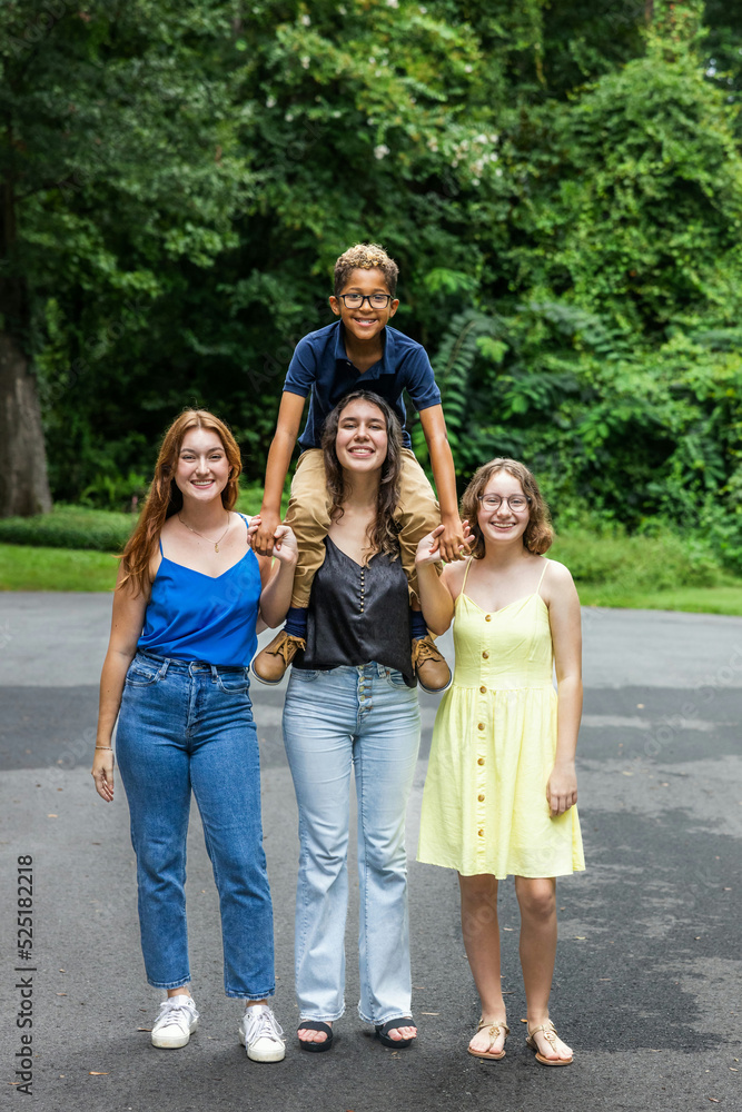 Four siblings with the youngest one sitting on his big sister's shoulders outside in the summer Four siblings with the youngest one sitting on his big sister's shoulders outside in the summer
