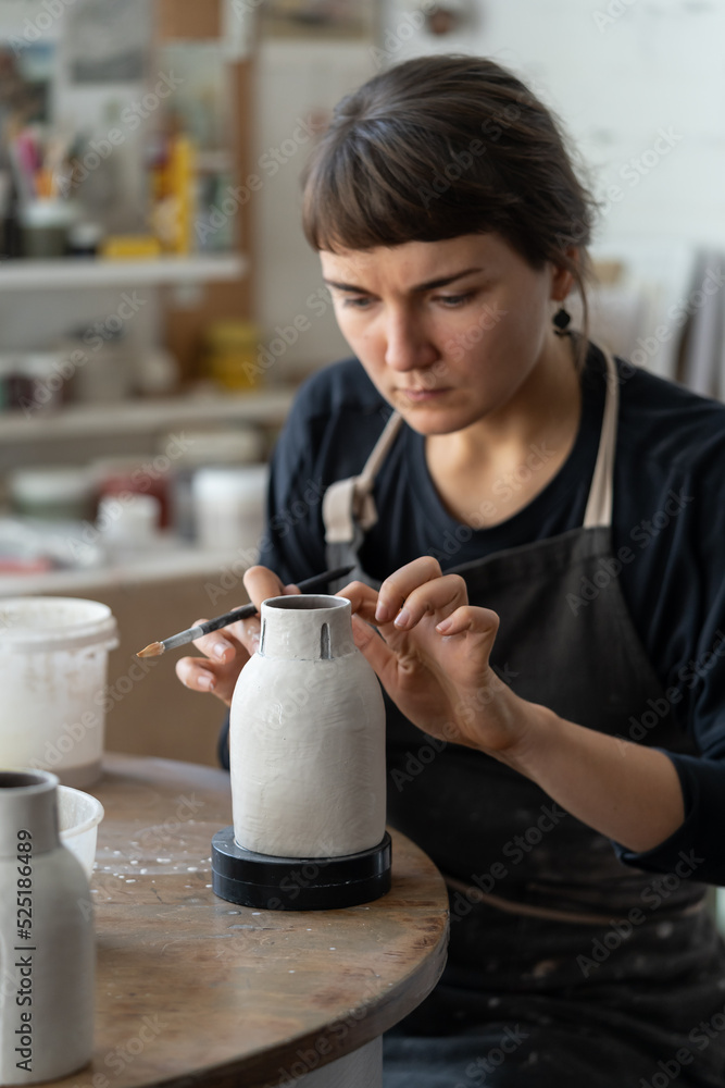 Stockfoto med beskrivningen Young female artisan in black apron works with  crockery sitting at round table in workshop against bright window. Brunette  woman enjoys painting handmade ceramic vase in pottery craft studio