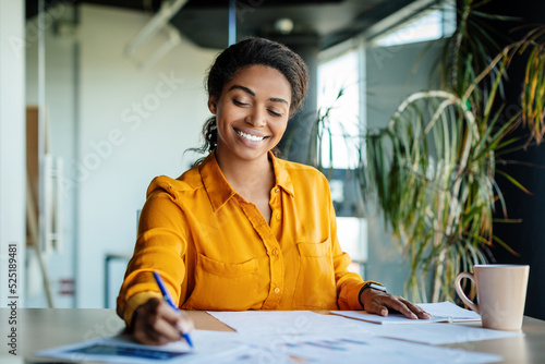 Happy black female company worker working with documents, writing and doing paperwork, sitting at workplace in office photo