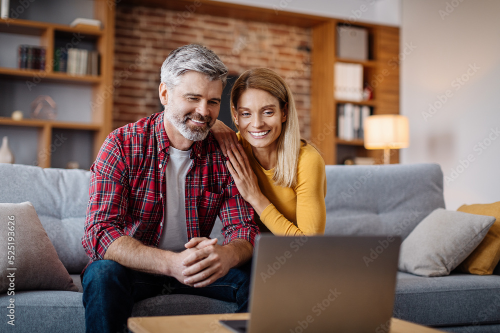 Cheerful middle aged caucasian man and woman watch video in laptop, making online call in living room