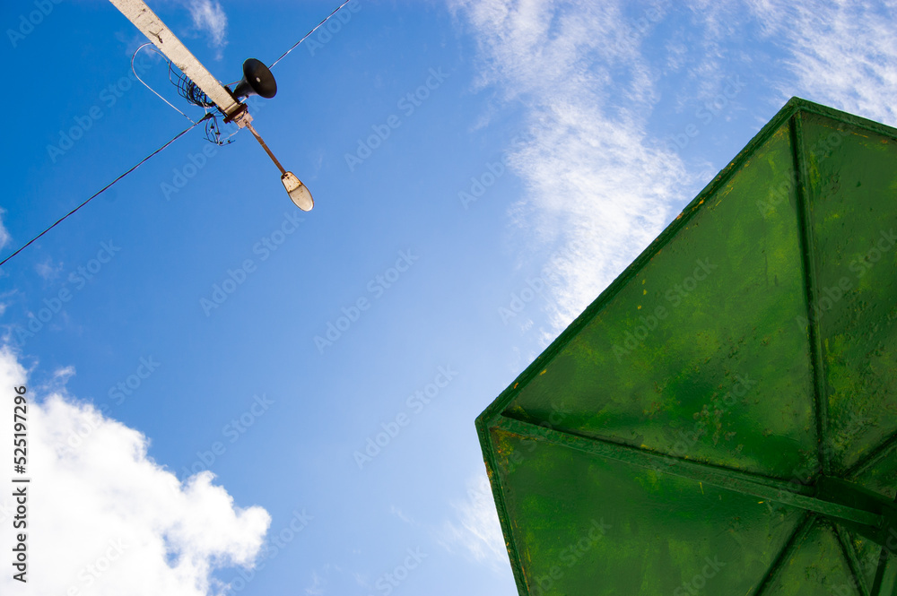 A green metal umbrella and a lantern against a blue sky with clouds. Bottom view A green metal umbrella and a lantern against a blue sky with clouds. Bottom view