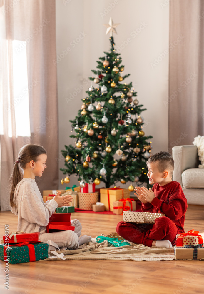 christmas, winter holidays and childhood concept - happy girl and boy in pajamas opening gifts sitting on floor in front of each other at home christmas, winter holidays and childhood concept - happy girl and boy in pajamas opening gifts sitting on floor in front of each other at home