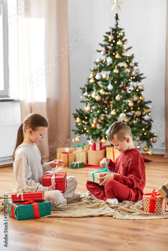 christmas  winter holidays and childhood concept - happy girl and boy in pajamas opening gifts sitting on floor in front of each other at home
