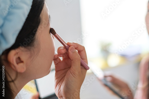 Asian woman doing makeup with brush in front of bathroom mirror.