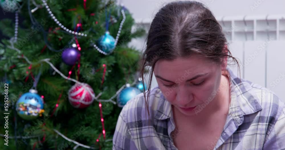 Exhausted disheveled brunette young woman in plaid shirt looks down intently, sitting next to a decorated Christmas tree, front view. She was tired of the festive fuss Exhausted disheveled brunette young woman in plaid shirt looks down intently, sitting next to a decorated Christmas tree, front view. She was tired of the festive fuss