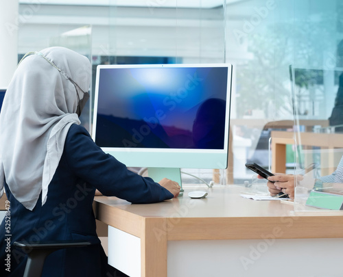 Back view of muslim lady using desktop computer sitting at desk office. Muslim woman customer service staff wearing mask working in office, serving customer. Covid-19 health protocol implementation.