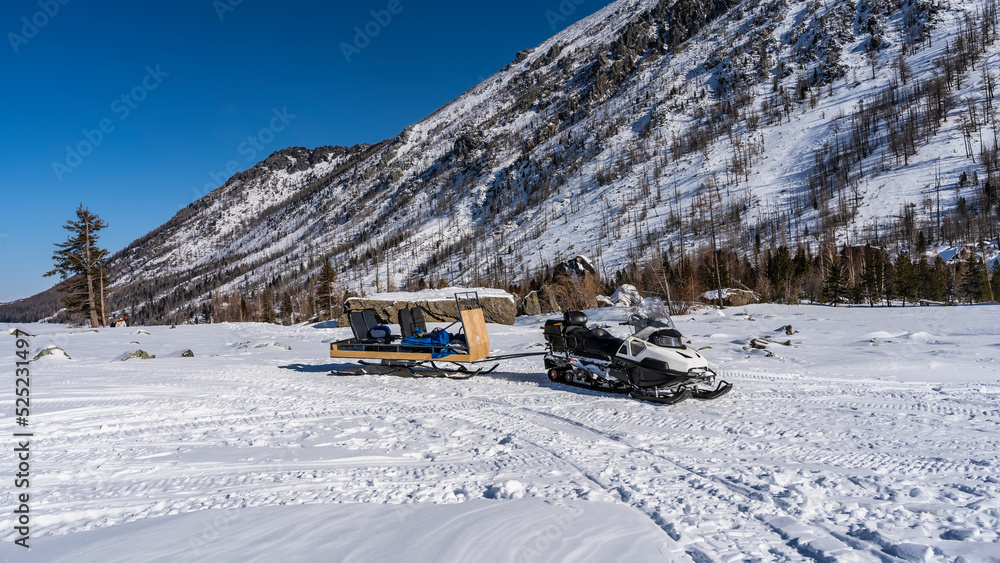 In a snowy valley there is a snowmobile with a sled attached. Traces of runners around. Forest on the mountainside. Clear blue sky. Altai In a snowy valley there is a snowmobile with a sled attached. Traces of runners around. Forest on the mountainside. Clear blue sky. Altai