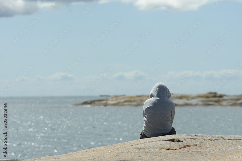 A lonely boy in a grey hoodie sits on a rock looking out over the sea A lonely boy in a grey hoodie sits on a rock looking out over the sea