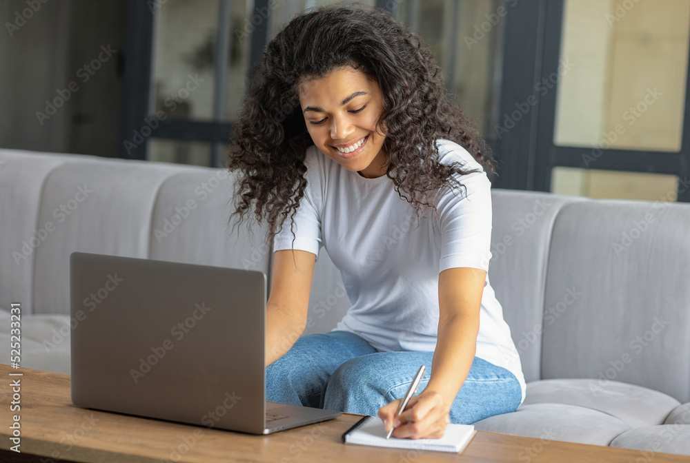 Online learning, distance education concept. Happy African American woman student or freelancer working or watching webinar using laptop, taking notes in notebook sitting on sofa at home