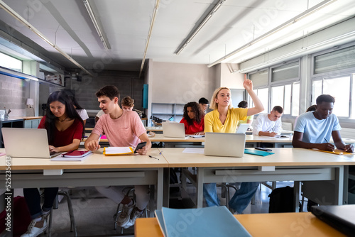 Teen female student raising arm to ask question about lesson in classroom.