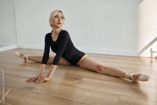 Ballerina in a black suit and pointe shoes doing stretching while sitting on the floor in a ballet studio