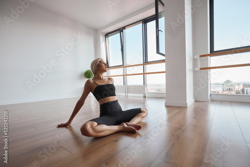 Young satisfied girl practicing choreographic movement on the floor. Attractive fit dancer stretching in the dance hall, bright ballet studio