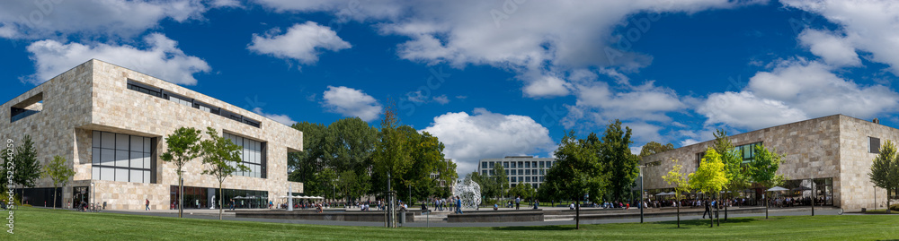 Moderne, quaderförmige Gebäude auf einem Gelände in einer Panoramaaufnahme mit grüner Wiese und aufgelockerter Bewölkung Moderne, quaderförmige Gebäude auf einem Gelände in einer Panoramaaufnahme mit grüner Wiese und aufgelockerter Bewölkung