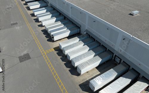 An aerial view above white trailers parked at a large warehouse, loading docks. Seen on a sunny day from a high, angled view.