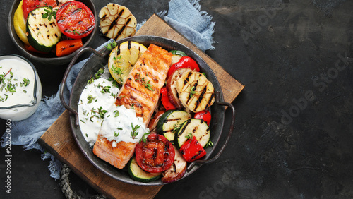 Salmon steak with vegetables and white sauce on dark background.