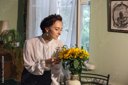 A stylish Ukrainian woman in vintage clothes, a black skirt and a white shirt, stands by the window in a cozy apartment with retro furniture. There is a vase with sunflowers on the table.