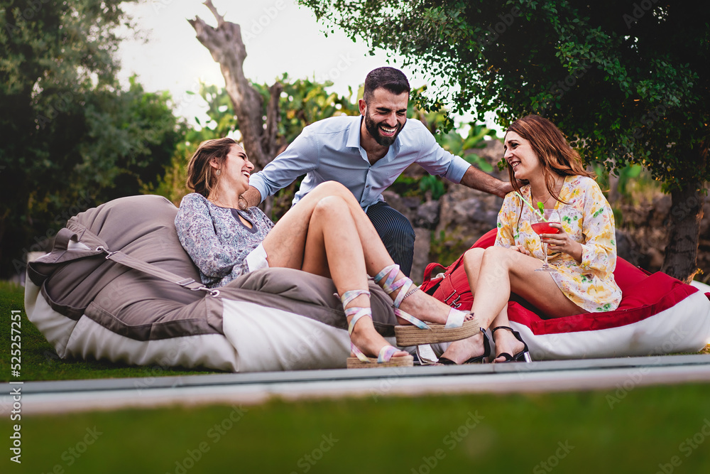 One young man and two women talking and laughing carefree sitting on bean bags at luxury backyard party drinking fancy cocktails