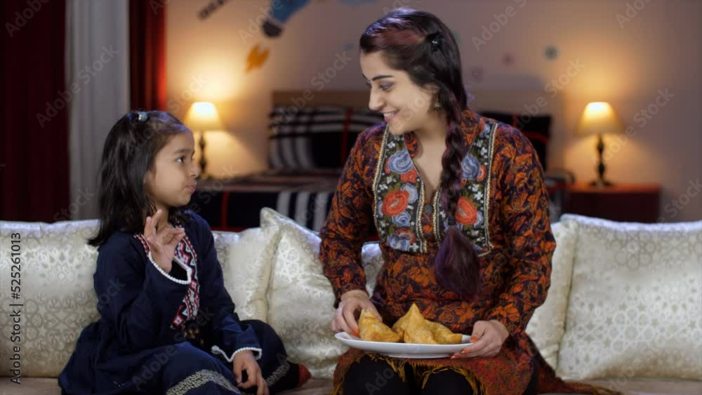 A young Indian mother feeding Samosa to her daughter - street food stuffed Samosa junk food home made. A little girl and her mother bingeing on unhealthy food - cheat days poor eating habits d...
