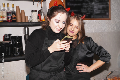 Halloween party at coffee shop, cafe. Funny, laughing, having fun, surprised women waiters wearing on Halloween costume, looking at smartphone. Halloween bakery. Young entrepreneur conceptual.
