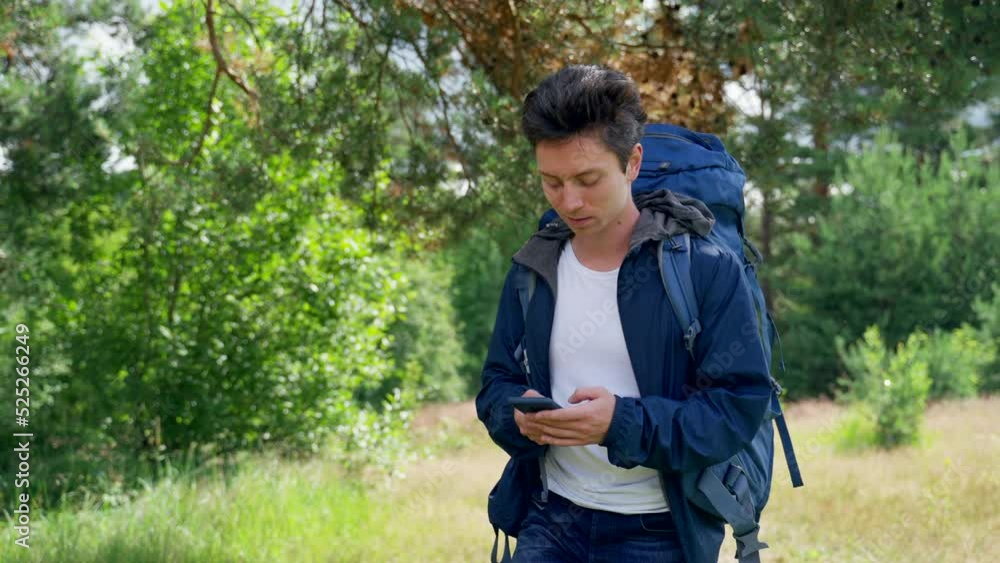 Black-haired guy hiker walks on meadow smiling and looking at phone for finding destination. Young man with blue backpack prefers hiking in forest Black-haired guy hiker walks on meadow smiling and looking at phone for finding destination. Young man with blue backpack prefers hiking in forest