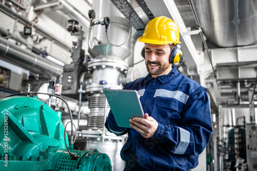 Industrial engineer standing by gas generator in power plant and controlling electricity production on his tablet.