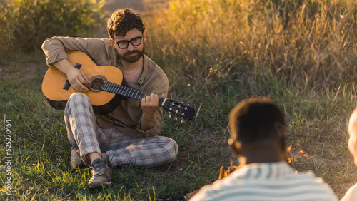 A young bearded caucasian man playing the guitar during a picnic in nature with his friends. People chilling near the fire, enjoying and watching the sunset. High quality photo