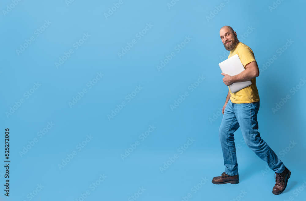 Full length body size view of attractive bearded smile middle-aged man going using laptop over bright blue color background. Casual clothing , yellow t-shirt and blue jeans. Full length body size view of attractive bearded smile middle-aged man going using laptop over bright blue color background. Casual clothing , yellow t-shirt and blue jeans.