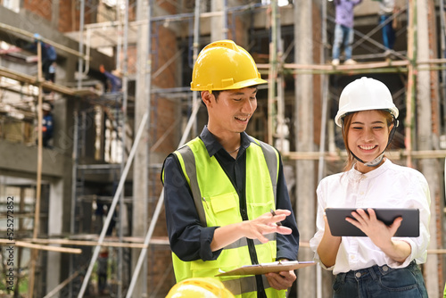 Smiling male supervisor and female investor wearing hardhats and reflective jackets inspecting commercial building construction site