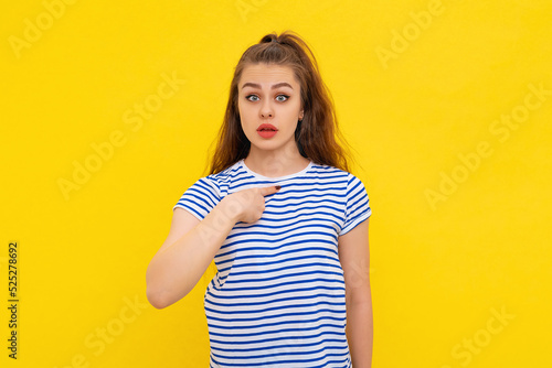 Shocked and worried young brunette woman pointing fingers at herself, frowning and sulking upset, standing in white-blue striped t shirt over yellow background