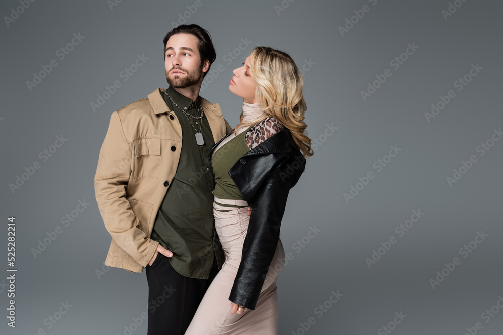 blonde woman in black leather jacket looking at man posing with hand in pocket isolated on grey