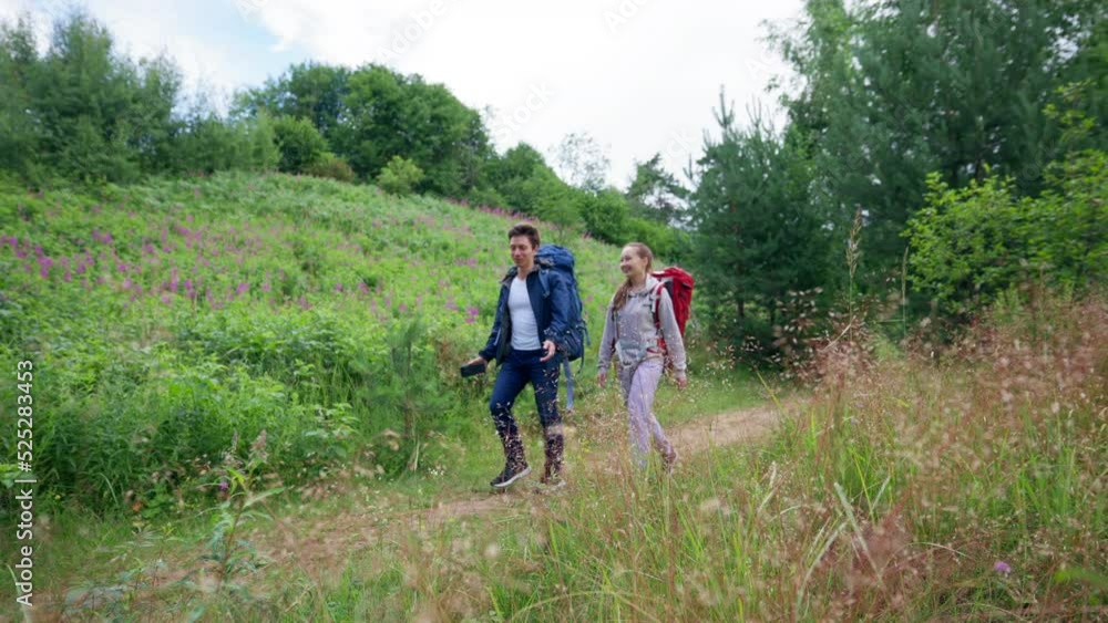 Couple of young tourists walks on footpath trying to find perfect spot for camping. Man and woman enjoy hiking walking past meadow with smile slow motion Couple of young tourists walks on footpath trying to find perfect spot for camping. Man and woman enjoy hiking walking past meadow with smile slow motion