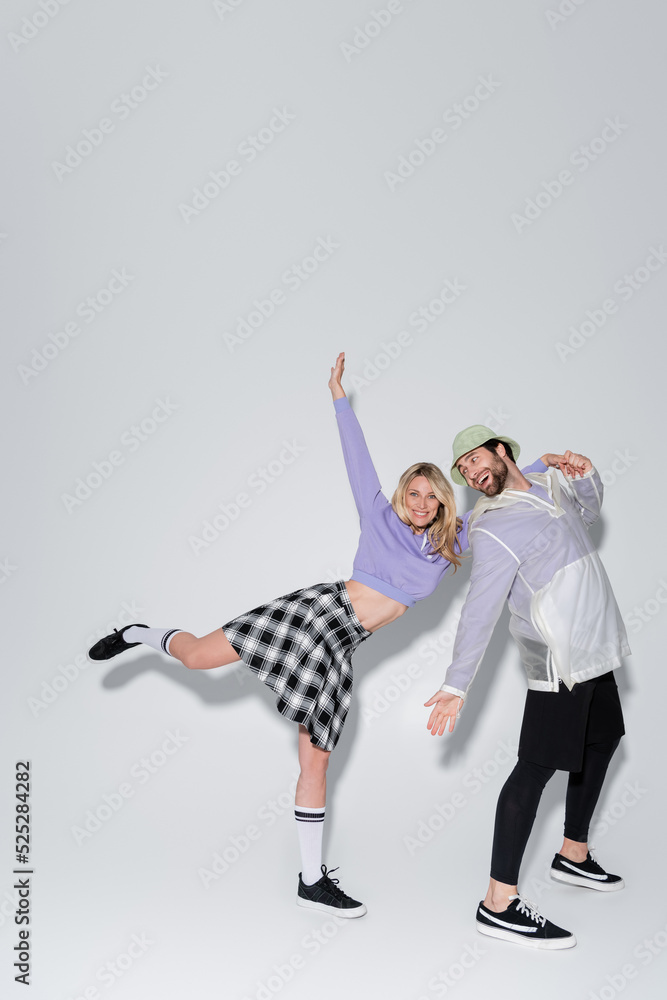 full length of cheerful woman in tartan skirt and longs socks with sneakers leaning on man in panama hat on grey