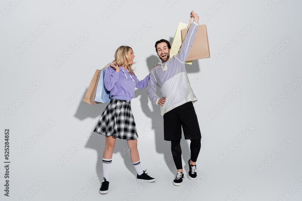 Naklejka premium full length of excited man and cheerful woman in tartan skirt holding shopping bags on grey