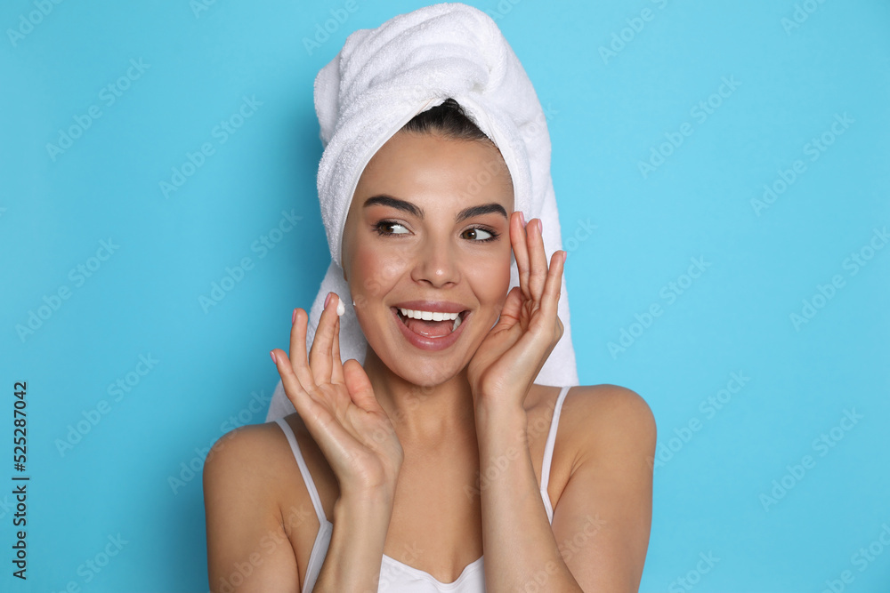 Beautiful young woman with towel applying cream on face against light blue background Beautiful young woman with towel applying cream on face against light blue background