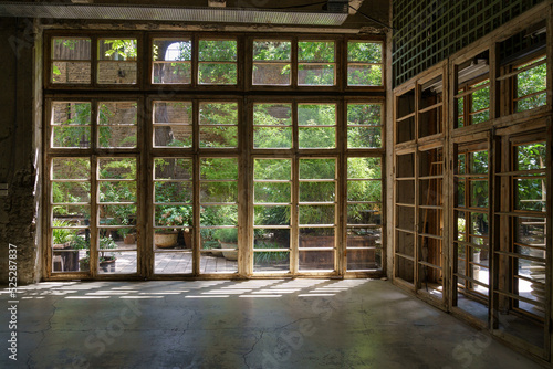 Interior of cottage house in process of renovation in countryside on sunny summer day. Sunlight breaks through big wooden frame of panoramic window overlooking garden with lush green plants and bushes © DimaBerlin
