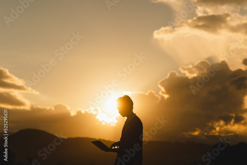 Silhouette of Asian farmer holding a laptop in the field examining crops at sunset.High technology agricultural occupation concept.