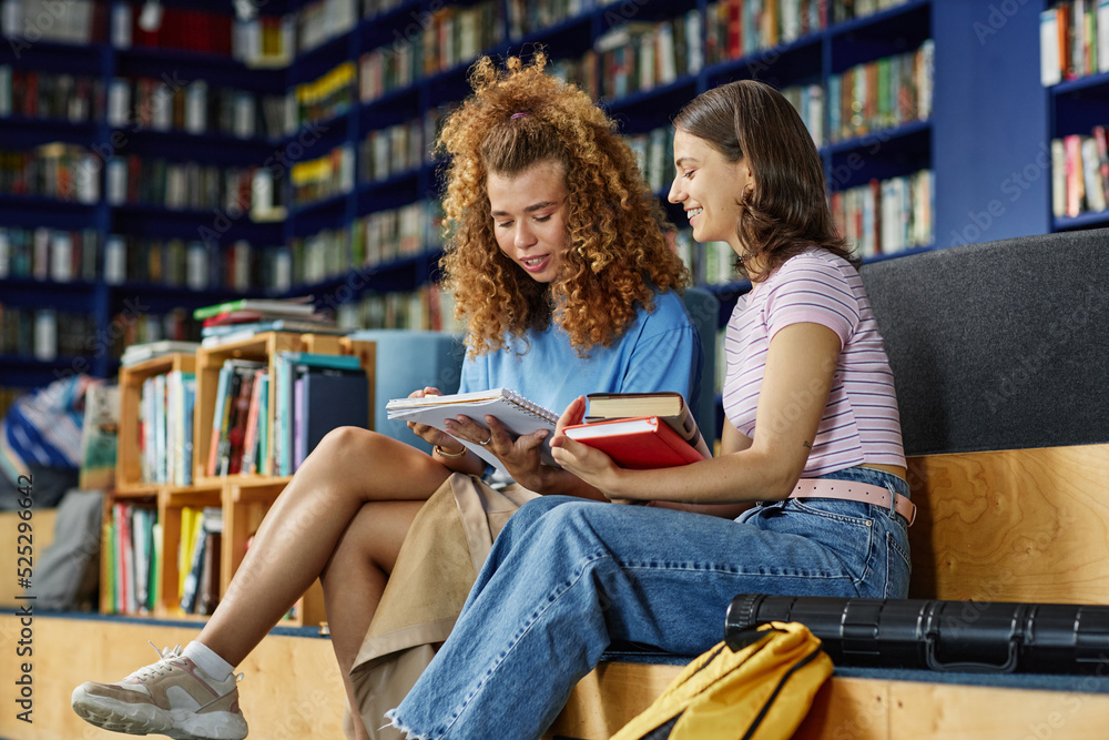 © Seventyfour - Vibrant portrait of two young girls reading books in college library and smiling, copy space