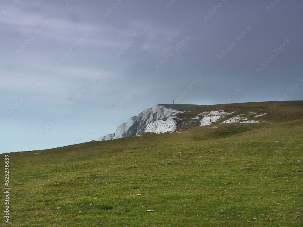 A sweep of cliff top heathland wilderness, topped by a distant monument, curves back on itself to reveal rugged chalk cliff under a storm clouded sky. A sweep of cliff top heathland wilderness, topped by a distant monument, curves back on itself to reveal rugged chalk cliff under a storm clouded sky.