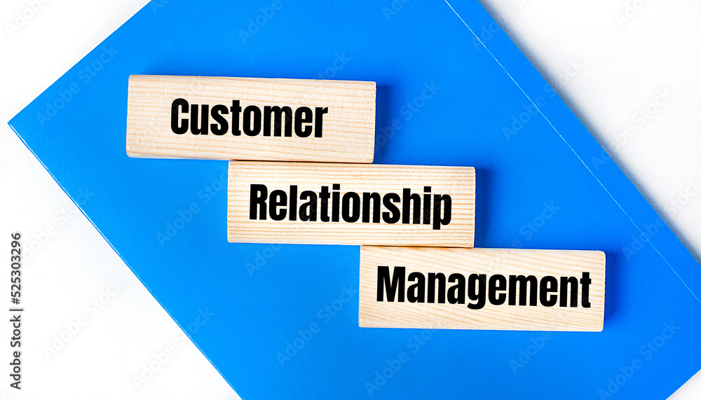 There is a blue notebook on a light gray background. Above are three wooden blocks with the words CRM Customer Relationship Management There is a blue notebook on a light gray background. Above are three wooden blocks with the words CRM Customer Relationship Management