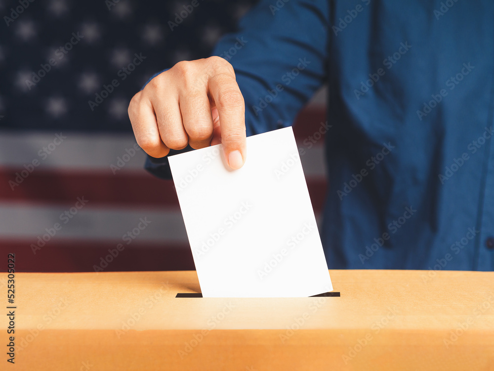 Hand voter holding ballot paper putting into the voting box at place election with an American flag background Hand voter holding ballot paper putting into the voting box at place election with an American flag background
