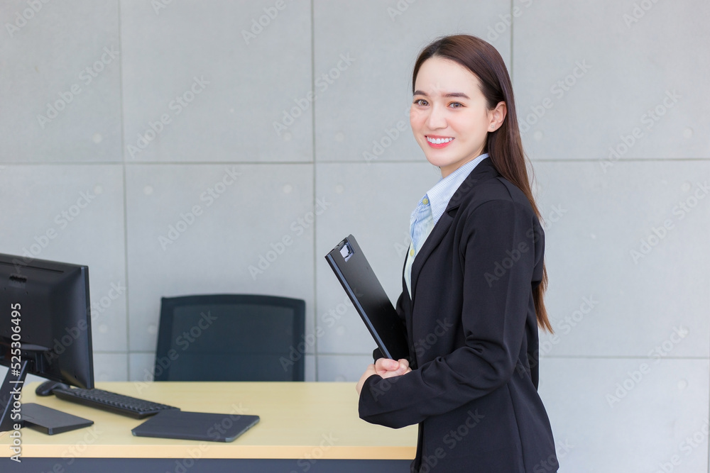 Young Asian professional working woman in a black suit holds clipboard in her hands and confident smiles in office room. Young Asian professional working woman in a black suit holds clipboard in her hands and confident smiles in office room.