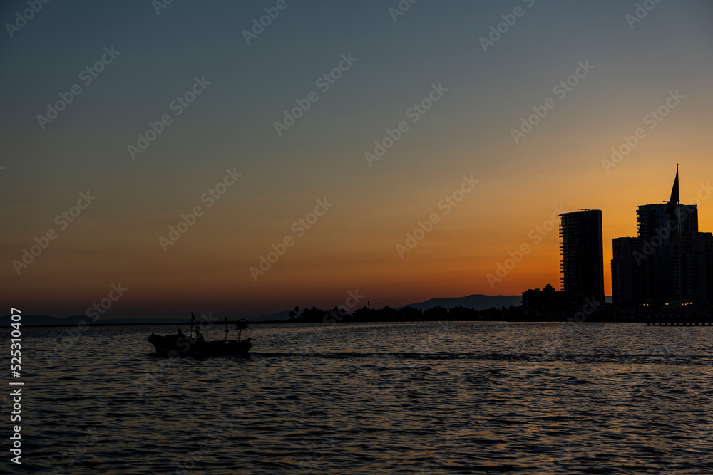 Fototapeta premium View of fishing boat with two fisherman gliding in the sea at sunset in Mavisehir, Karsiyaka, İzmir. Beautiful sunlight over skyline. Selective focus, noise effect and grainy texture.