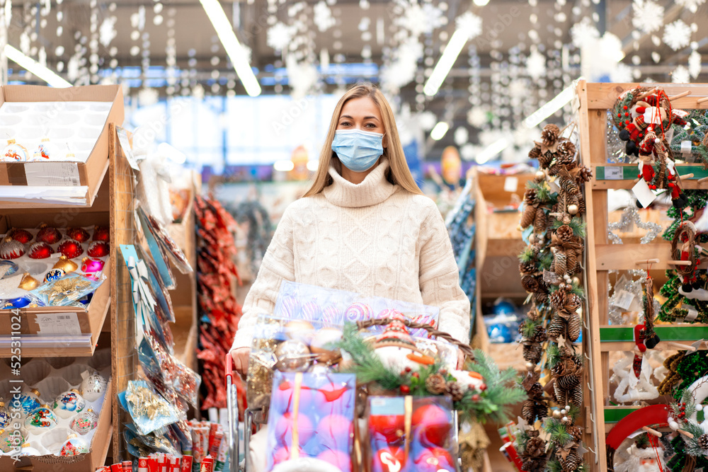 Christmas shopping. Woman with cart shopping in protective medical mask watching Christmas ornament in shopping mall on Holiday sale.