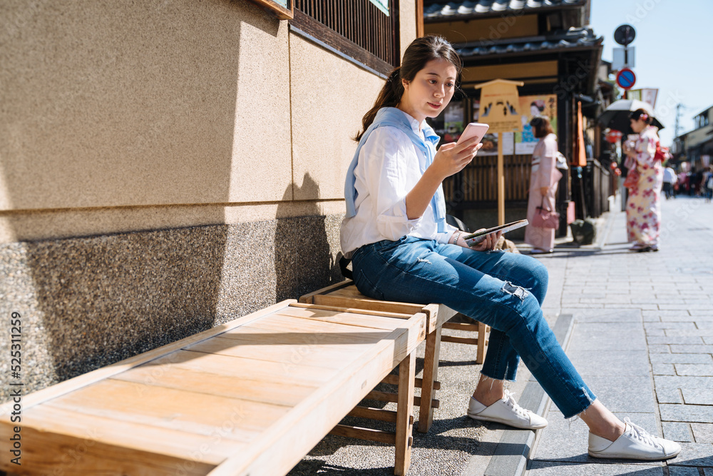 asian girl in casual wear is checking phone messages while taking a rest sit on the wooden bench at hanamikoji street in gion district, Kyoto japan with sunlight